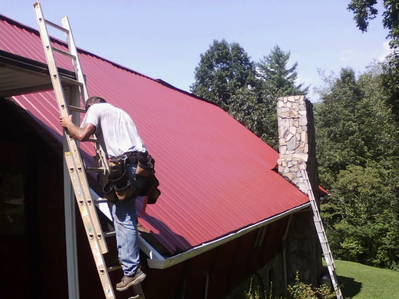 Roofer on a ladder beside a red metal roof and stone chimney — Soffit & Fascia Repair by Roofing Brothers in Alva